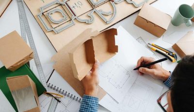 Box maker looking at box, drawing it’s schematics Box maker working behind a table in a private workshop, drawing scematics with a pencil, tools scattered across. Rulers, paper knife and special board with a cutting form.